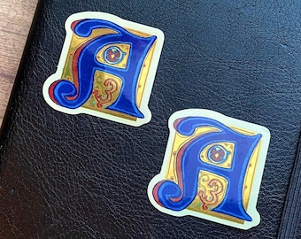 Könnte beinhalten: Zwei goldene und blaue Aufkleber mit dem Buchstaben A und komplizierten Designs. Die Aufkleber befinden sich auf einer schwarzen Lederoberfläche.