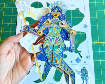 Könnte beinhalten: Eine detaillierte Papierpuppe einer Ozeanprinzessin, mit einem blau-goldenen Farbschema. Die Puppe hält eine Fahne und ist mit sternförmigen Akzenten verziert. Der Text "Ocean Princess Paper Doll" ist sichtbar.