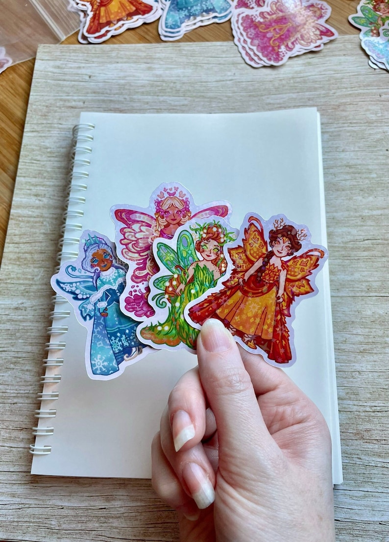 May include: A collection of colorful fairy stickers with intricate designs. The stickers feature fairies in various outfits and poses, with details like wings, crowns, and floral accents. The stickers are held over a white spiral notebook.