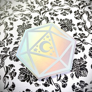 May include: A holographic iridescent sticker of a white 20-sided die with a crescent moon and stars inside. The sticker is on a white background with a black floral pattern.