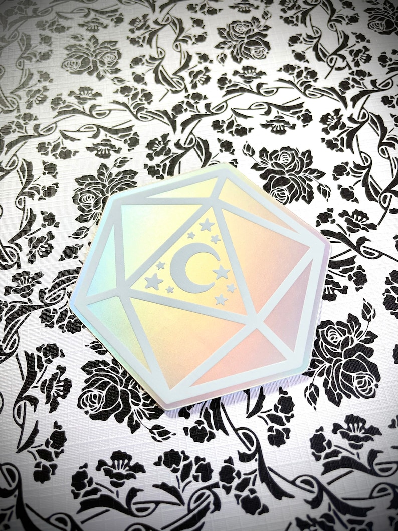 May include: A holographic iridescent sticker of a white 20-sided die with a crescent moon and stars inside. The sticker is on a white background with a black floral pattern.