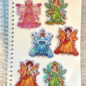 May include: A collection of six fairy stickers on a white spiral notebook. The stickers feature detailed illustrations of fairies in various outfits and colors, including pink, green, gold, and blue, with iridescent accents. The artist's name, "Hali Jensen Art," is visible.