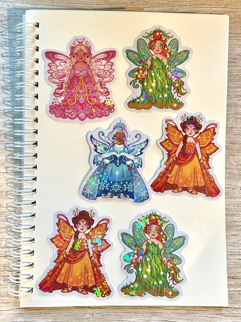 May include: A collection of six fairy stickers on a white spiral notebook. The stickers feature detailed illustrations of fairies in various outfits and colors, including pink, green, gold, and blue, with iridescent accents. The artist's name, "Hali Jensen Art," is visible.