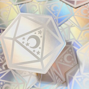 May include: A holographic iridescent sticker of a white, geometric, 12-sided die with a crescent moon and stars inside. The sticker is surrounded by other holographic stickers.