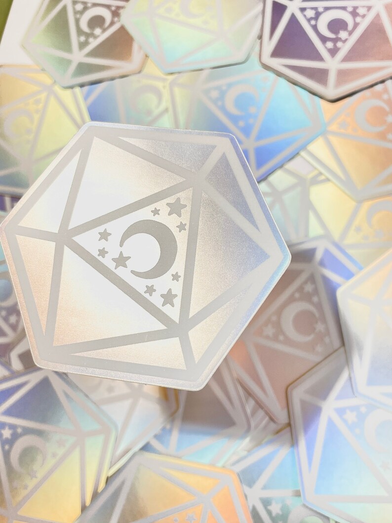 May include: A holographic iridescent sticker of a white, geometric, 12-sided die with a crescent moon and stars inside. The sticker is surrounded by other holographic stickers.