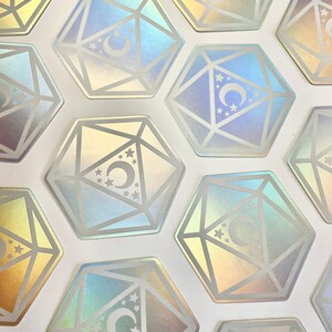 May include: A set of iridescent holographic stickers featuring a crescent moon and stars inside a geometric design resembling a twenty-sided die.