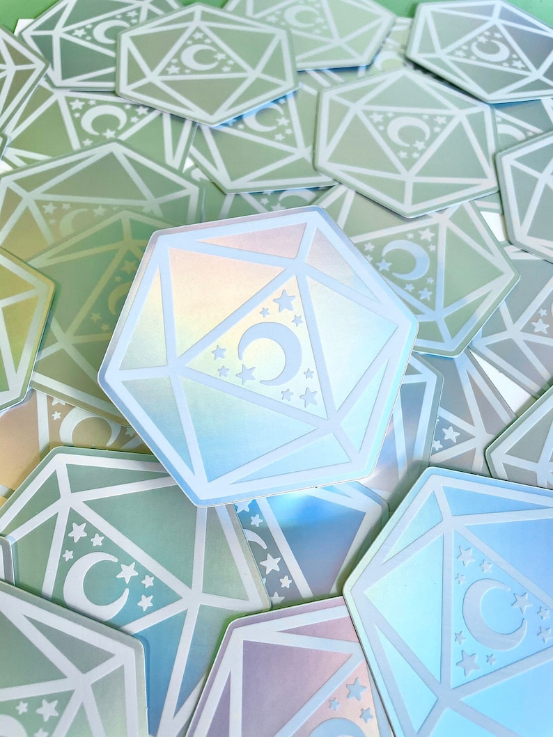 May include: A holographic iridescent sticker of a 20-sided die with a crescent moon and stars. The sticker is white with a green and blue iridescent background.