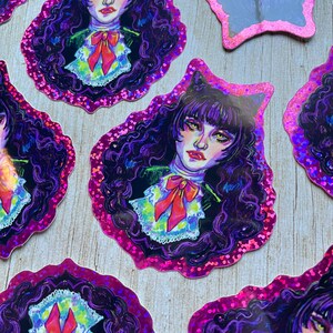 K&ouml;nnte beinhalten: Ein Satz holografischer Aufkleber mit einer Comicfigur mit schwarzen Haaren, violetten Augen und einer roten Schleife. Die Aufkleber befinden sich auf einem rosa Hintergrund.
