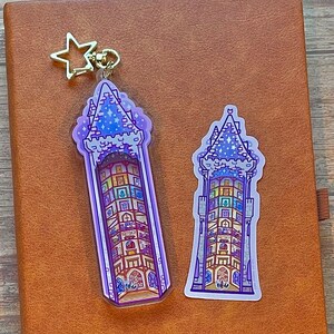 May include: Two purple and blue tower-shaped keychains with a gold star charm. The keychain on the left is larger and has a clear background. The keychain on the right is smaller and has a white background.