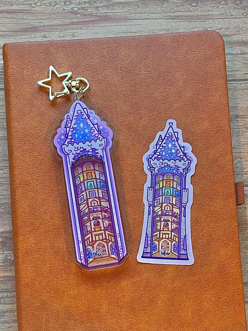May include: Two purple and blue tower-shaped keychains with a gold star charm. The keychain on the left is larger and has a clear background. The keychain on the right is smaller and has a white background.