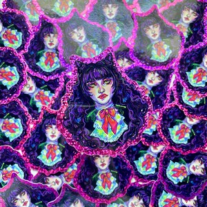 K&ouml;nnte beinhalten: Ein Stapel Aufkleber mit einem Comic-Charakter mit schwarzen Haaren, violetten Augen und einer rosa Schleife. Der Charakter tr&auml;gt einen wei&szlig;en Kragen und ein schwarzes Kleid. Die Aufkleber haben einen rosa Glitzer-Rand.