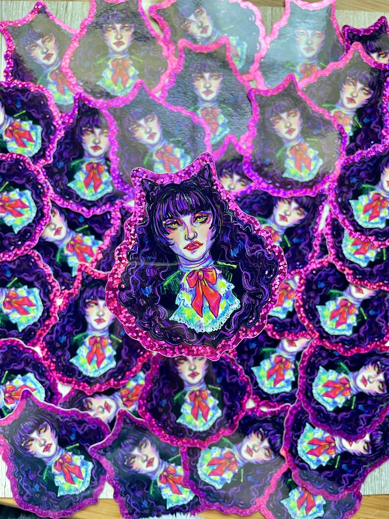 K&ouml;nnte beinhalten: Ein Stapel Aufkleber mit einem Comic-Charakter mit schwarzen Haaren, violetten Augen und einer rosa Schleife. Der Charakter tr&auml;gt einen wei&szlig;en Kragen und ein schwarzes Kleid. Die Aufkleber haben einen rosa Glitzer-Rand.