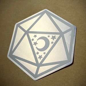 May include: A silver metal d20 dice with a crescent moon and stars design. The dice is a popular game piece used in tabletop role-playing games.