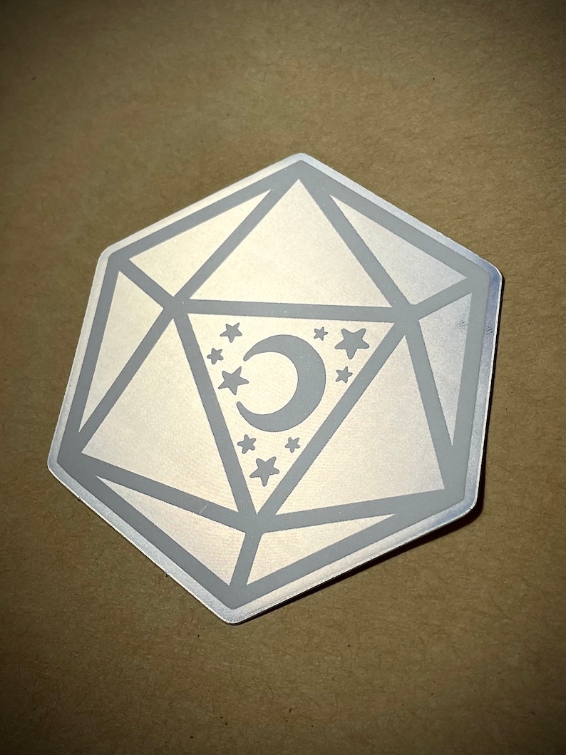 May include: A silver metal d20 dice with a crescent moon and stars design. The dice is a popular game piece used in tabletop role-playing games.