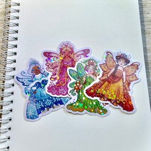 May include: Four colorful fairy stickers with iridescent accents. Each fairy is depicted in a unique outfit, including a winter-themed blue dress, a pink gown, a green dress, and a golden dress. The stickers are placed on a white spiral-bound notebook.