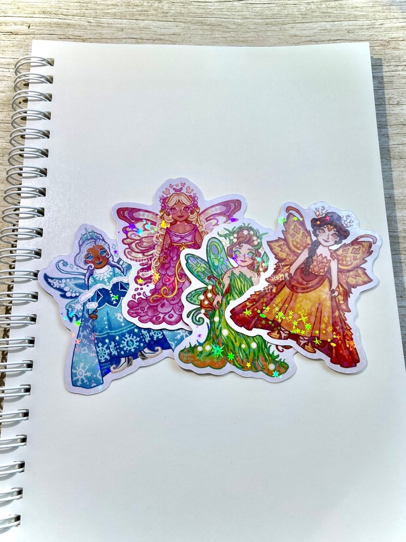 May include: Four colorful fairy stickers with iridescent accents. Each fairy is depicted in a unique outfit, including a winter-themed blue dress, a pink gown, a green dress, and a golden dress. The stickers are placed on a white spiral-bound notebook.