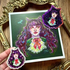 K&ouml;nnte beinhalten: Eine violette und gr&uuml;ne Illustration einer Frau mit Katzenohren und einer Schleife. Die Frau ist von einem gr&uuml;n-violetten Nebel umgeben. Die Illustration ist auf einer wei&szlig;en Karte gedruckt und in einem goldenen Rahmen gerahmt. Es gibt zwei kleinere Versionen der Illustration auf Aufklebern.