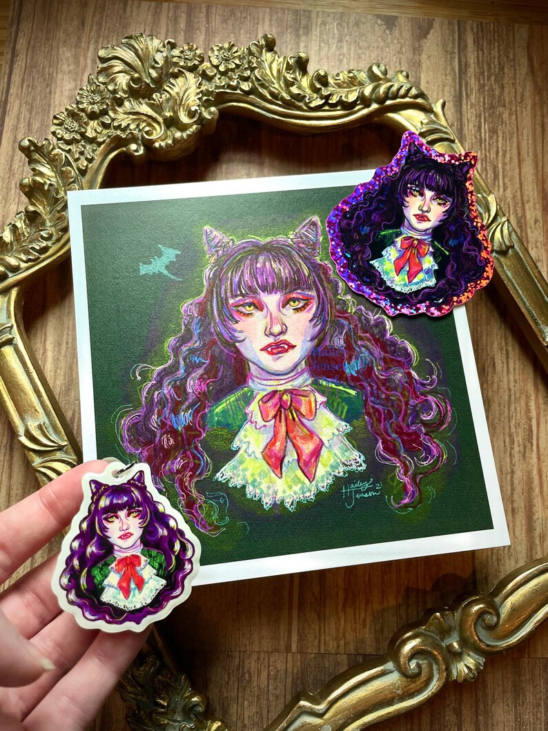 K&ouml;nnte beinhalten: Eine violette und gr&uuml;ne Illustration einer Frau mit Katzenohren und einer Schleife. Die Frau ist von einem gr&uuml;n-violetten Nebel umgeben. Die Illustration ist auf einer wei&szlig;en Karte gedruckt und in einem goldenen Rahmen gerahmt. Es gibt zwei kleinere Versionen der Illustration auf Aufklebern.