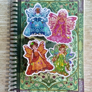 May include: A spiral-bound notebook with four fairy stickers. The stickers depict fairies in winter, spring, summer, and autumn attire. The notebook has a green floral design and a purple border.