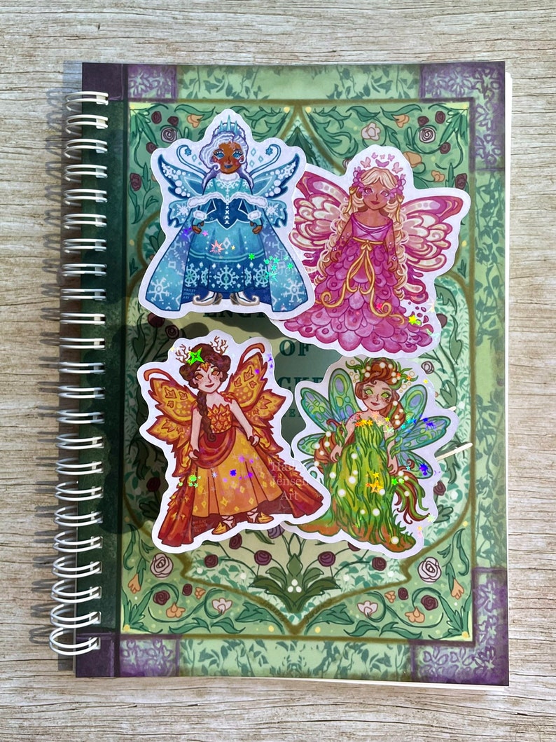 May include: A spiral-bound notebook with four fairy stickers. The stickers depict fairies in winter, spring, summer, and autumn attire. The notebook has a green floral design and a purple border.
