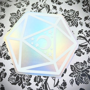 May include: A white iridescent sticker with a holographic finish, shaped like a twenty-sided die with a crescent moon and stars inside.