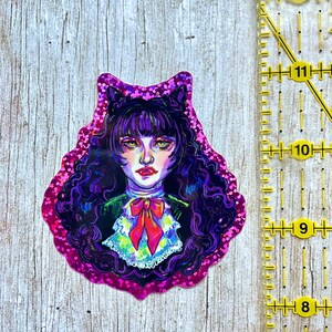 K&ouml;nnte beinhalten: Ein holografischer Aufkleber mit einer Cartoon-Illustration einer Frau mit langen schwarzen Haaren, die ein schwarz-wei&szlig;es Kleid mit einer roten Schleife tr&auml;gt. Der Hintergrund ist ein rosa-violetter Wirbel.