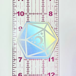 May include: A holographic iridescent sticker of a white dice with a crescent moon and stars inside. The sticker is approximately 1 inch in diameter.