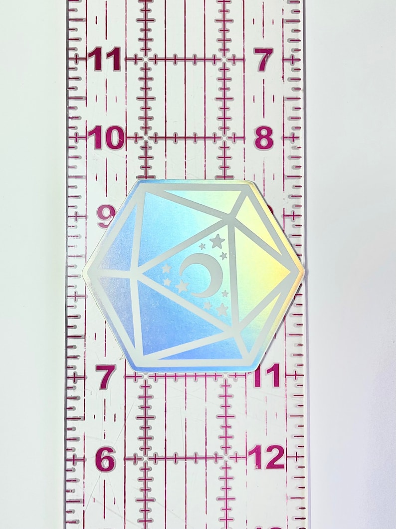 May include: A holographic iridescent sticker of a white dice with a crescent moon and stars inside. The sticker is approximately 1 inch in diameter.