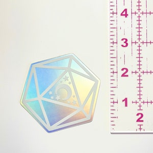 May include: A holographic iridescent sticker of a 20-sided die with a crescent moon and stars inside. The die is approximately 2 inches in diameter.