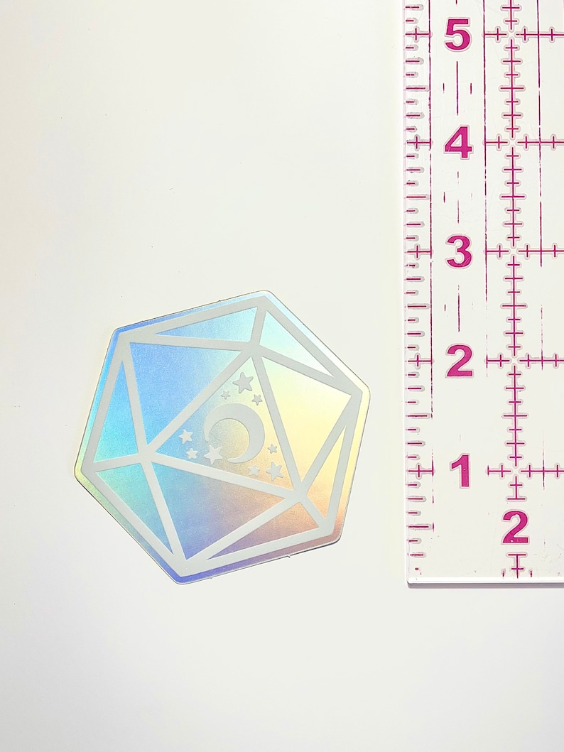 May include: A holographic iridescent sticker of a 20-sided die with a crescent moon and stars inside. The die is approximately 2 inches in diameter.
