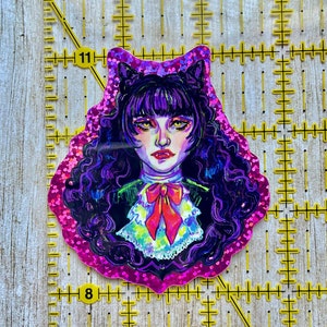 K&ouml;nnte beinhalten: Ein Aufkleber mit einer Cartoon-Illustration einer Frau mit lila Haaren und Katzenohren. Sie tr&auml;gt ein schwarz-wei&szlig;es Kleid mit einer roten Schleife. Der Aufkleber hat einen pinkfarbenen Glitzer-Hintergrund.
