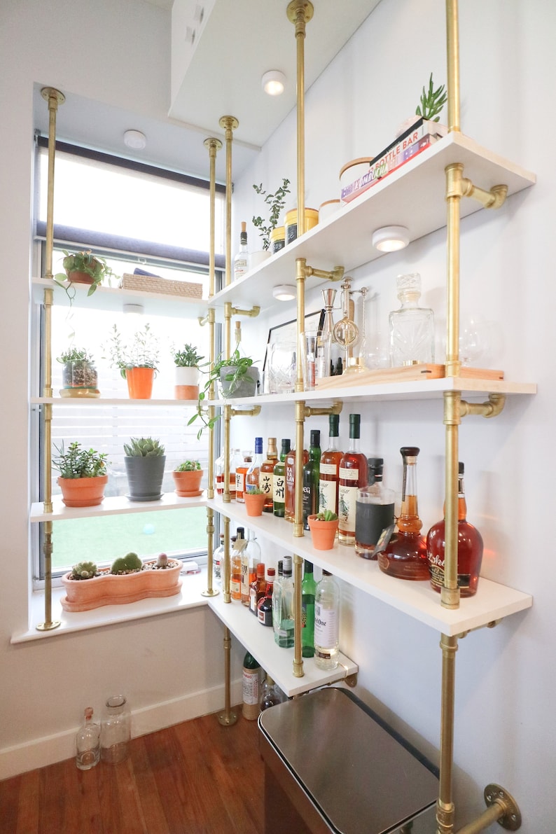 A large shelving unit made of brass hardware and white wood shelves is installed in the corner of a home. There are plants on one side in front of a window. The other side bar bottles and barware.