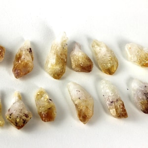 May include: A collection of raw citrine crystal points, varying in size and shape. The crystals display a range of colors from pale yellow to golden brown, with some having a translucent quality. The crystals are arranged on a white surface.