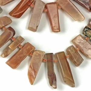 May include: A necklace of rectangular, polished gemstone beads in shades of tan, brown, and peach. The beads are strung on orange thread, creating a semi-circular arrangement. The gemstones have a natural, organic appearance.