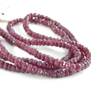 May include: Strands of faceted, deep red ruby beads. The beads are small and uniform in size, strung together to create a necklace or bracelet. The ruby beads have a polished, reflective surface, and are displayed against a white background.