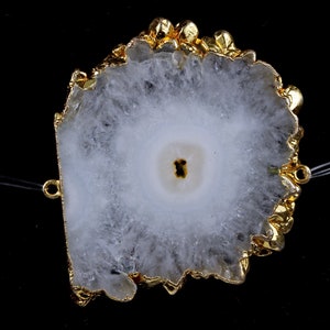 May include: A white and gray agate slice with a gold-plated edge. The stone has a central eye-like pattern and is strung with two thin, clear cords. The gold edge has an irregular, textured appearance.