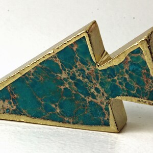 May include: A gold-plated pendant with a turquoise stone inlay. The pendant has a geometric arrow shape with a gold border. The turquoise stone has a marbled pattern with brown and beige veins. The pendant has a small gold loop for a chain.
