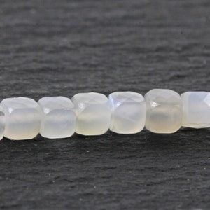 May include: A strand of pale, translucent, square-cut beads. The beads have a subtle, milky color and a faceted surface, creating a soft, luminous appearance. The beads are strung together, suggesting their use in jewelry or craft projects.