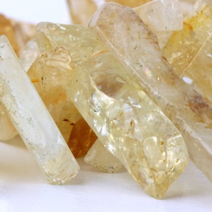 May include: Close-up of a pile of raw, natural citrine crystals. The crystals are a translucent yellow color with visible inclusions and varying shapes. The crystals are unpolished and have a rough texture.