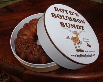 May include: A white round cake tin with a brown bundt cake inside. The lid of the tin is white with brown text that reads "Boyd's Bourbon Bundt" and a cartoon illustration of a Texas Longhorn steer. The text also includes "A Texas Original!" and the website "www.boydsbourbonbundt.com".