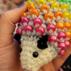 May include: A crocheted hedgehog with a rainbow of colorful beads for quills. The hedgehog has black eyes and a pink nose.