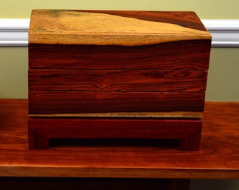 Puede incluir: Una caja de madera con un diseño en capas, que presenta un patrón de veta de madera natural y un acabado marrón rojizo. La caja está sentada sobre una mesa de madera.