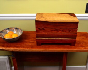 Puede incluir: Una caja de madera con un diseño en capas, que presenta un rico color marrón rojizo y intrincados patrones de veta de madera. La caja está sentada sobre una mesa de madera con un tazón de naranjas.