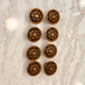 Puede incluir: Ocho tiradores de armario antiguos de bronce con diseño floral. Los tiradores son redondos con un centro en relieve y detalles decorativos en los bordes. Los tiradores están dispuestos en dos filas de cuatro.