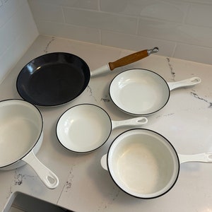 May include: A set of five cast iron cookware pieces, including a black frying pan, a white frying pan, and three white pots with black rims. The cookware is arranged on a white worktop.