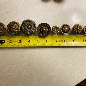May include: Nine antique brass cabinet knobs with intricate designs, measured on a yellow tape measure. The knobs are arranged in a row from left to right, with the tape measure running along the bottom.