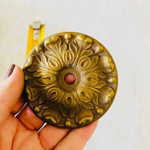 以下が含まれることがあります： 隆起した花柄と小さな赤い中心を持つ、丸い真鍮色の装飾的なアクセント。アクセントは、わずかに古びたアンティークな外観をしています。