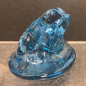 May include: A blue glass frog figurine. The frog is sitting on a round base. The glass is a translucent blue color. The frog has a textured surface.