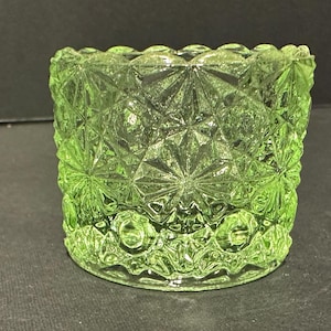 May include: A green glass votive candle holder with a geometric starburst pattern. The sides have a repeating diamond and star design. The bottom edge has a row of small circles. The glass is a light, translucent green.