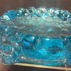May include: A turquoise glass turtle-shaped ashtray. The ashtray features a textured shell design and is made of transparent glass. The turtle has a head, four flippers, and a tail. The ashtray is sitting on a clear surface.